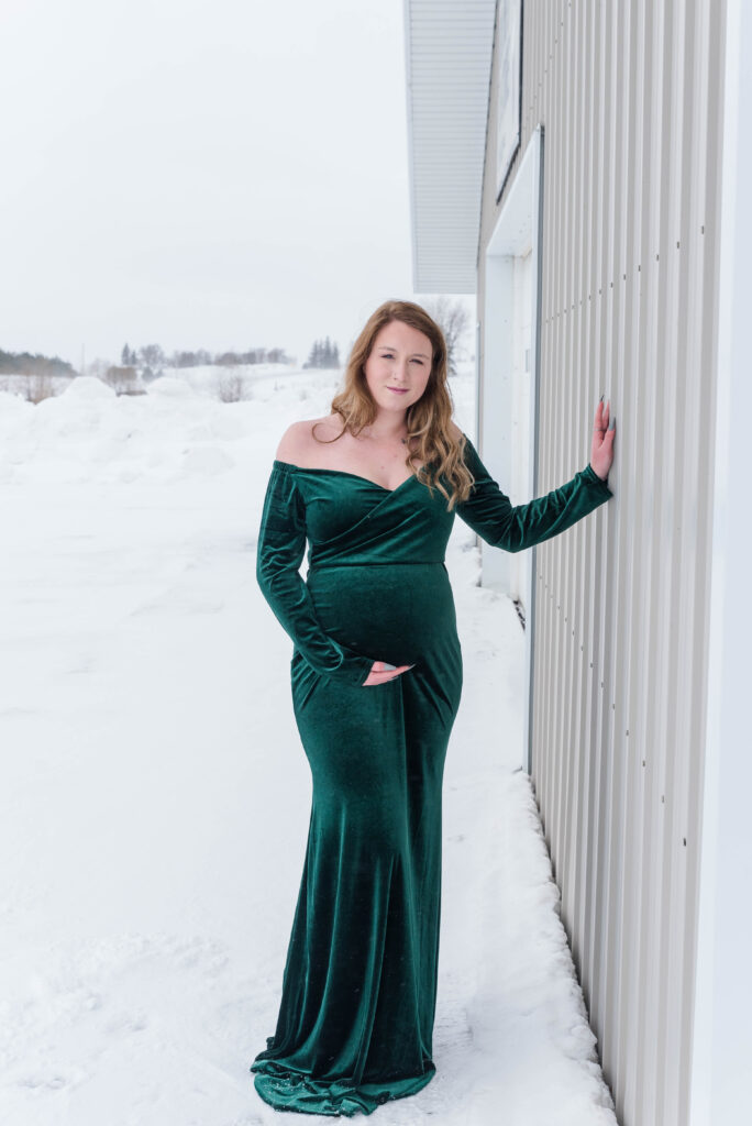 Pregnant Woman standing against metal siding with snow on the ground wearing an emerald green dress