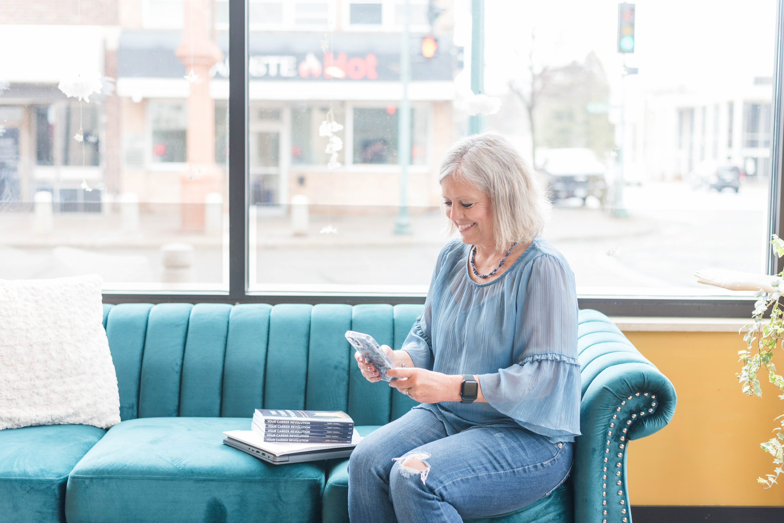 Woman wearing blue shirt sitting on teal couch holding phone.