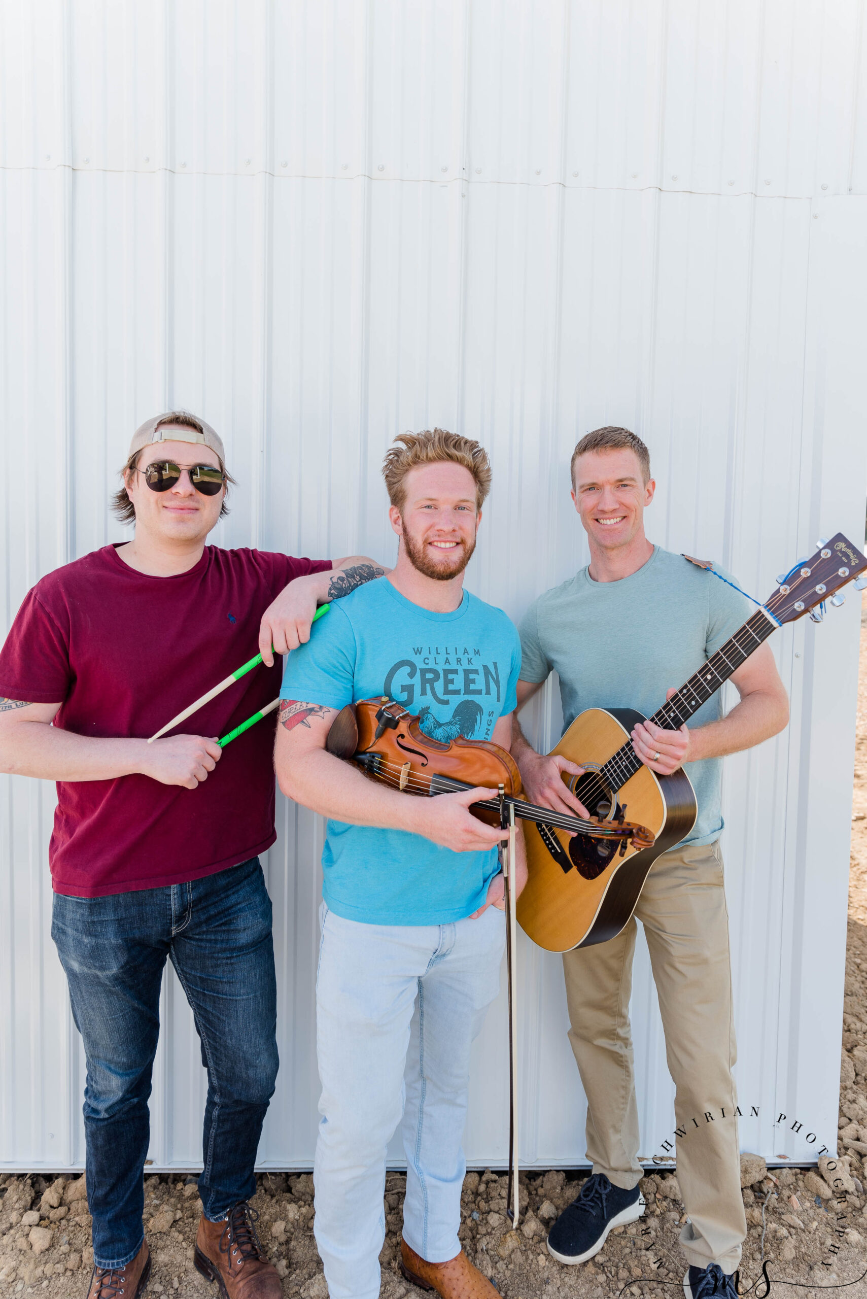 Three Band Members standing together, one holding his drum sticks another a violin and the third a guitar.