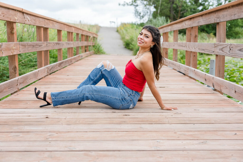 Senior Session in Vergas, MN. Girl sitting on a bridge wearing a red top, jeans and black heels 