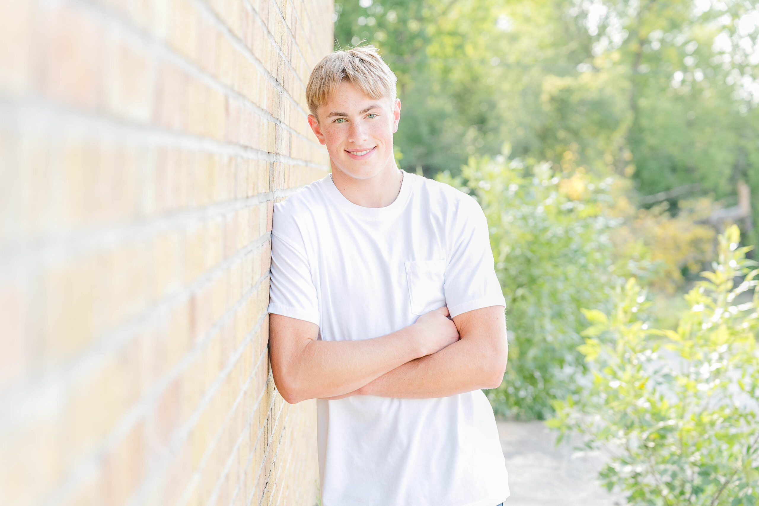 High School Senior wearing a white short sleeved shirt leaning up against a tan brick wall with green trees in the background